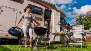 Een afbeelding van een vrouw die aan het barbecueën is op een camping met achter haar een kampeerwagen