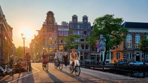 Een afbeelding van mensen die fietsen over een brug in Amsterdam met grachtenhuizen en een zonsondergang op de achtergrond