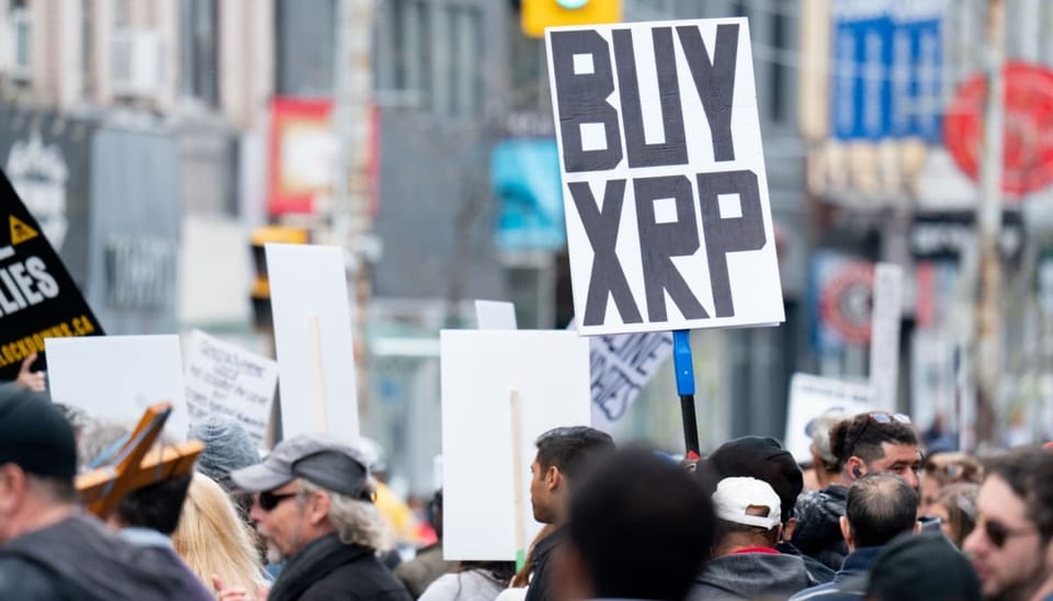 Een afbeelding van een menigte waarvan een aanwezige een bord met 'buy [de crypto] XRP'