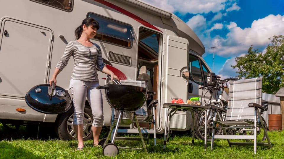 Een afbeelding van een vrouw die aan het barbecueën is op een camping met achter haar een kampeerwagen