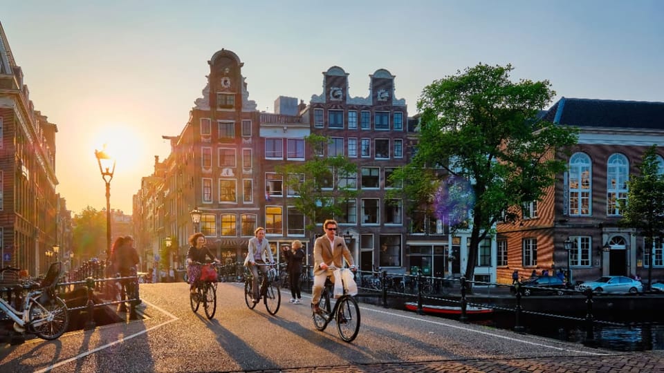 Een afbeelding van mensen die fietsen over een brug in Amsterdam met grachtenhuizen en een zonsondergang op de achtergrond