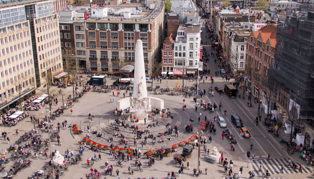 Een afbeelding van het plein van de Dam in Amsterdam, met daarbij het monument en een hoop Nederlanders